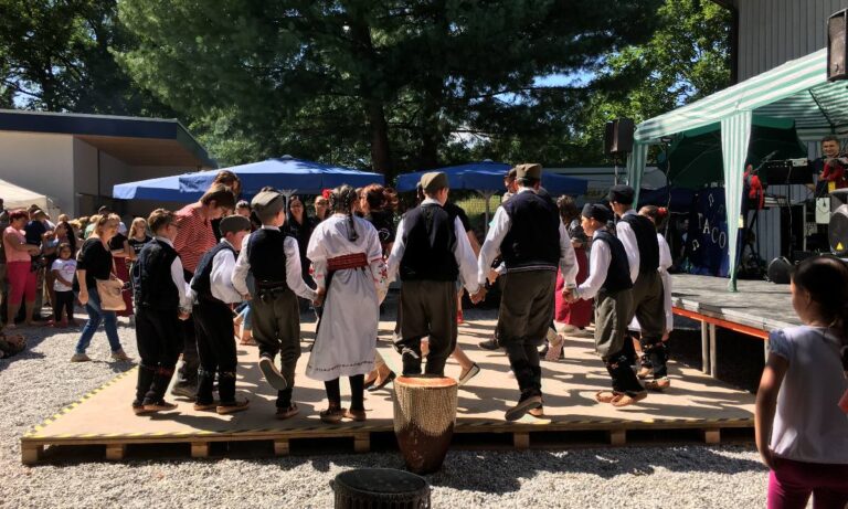 Menschen in traditioneller Kleidung tanzen in einer Gruppe auf einer Bühne im Freien bei sonnigem Wetter.