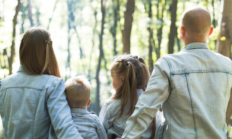 Eine Familie, bestehend aus Eltern und zwei Kindern, steht Rücken an Rücken und blickt in einen sonnigen Wald. Alle tragen Jeansjacken, was eine harmonische und familiäre Atmosphäre symbolisiert.