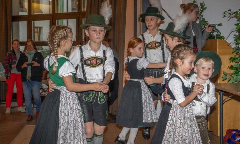 Eine Gruppe von Kindern in traditioneller bayerischer Tracht tanzt gemeinsam in einem festlich dekorierten Raum. Die Jungen tragen Lederhosen und grüne Hüte mit Federn, während die Mädchen Dirndl tragen. Im Hintergrund beobachten Erwachsene das Geschehen mit Interesse.