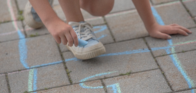 Eine Person hält kniend eine blaue Kreide in der Hand. Sie hat blaue Schuhe an und hat eine fünf auf den Boden gemalt.